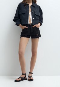Czarna krótka kurtka dżinsowa z srebrnymi guzikami, zestawiona z czarnymi, postrzępionymi krótkimi spodniami dżinsowymi oraz czarnymi, płaskimi sandałami.