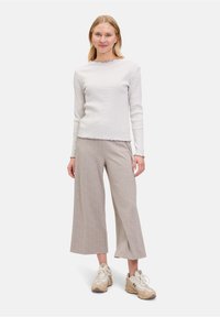 Gestreiftes Langarmoberteil mit Wellenmuster an den Säumen; kombiniert mit hellgrauen weit geschnittenen Hosen; cremefarbene Sneakers runden das Outfit ab. Der Hintergrund ist weiß.