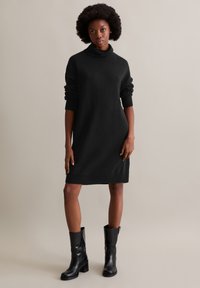 Schwarzes Strickkleid mit hohem Stehkragen und gerippten Bündchen, das eine gerade Silhouette und einen knielangen Saum hat. Getragen mit schwarzen High-Heel-Stiefeln.