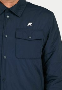 Giacca blu navy con bottoni, dotata di una tasca e di una toppa con logo. Presenta una consistenza liscia e un design classico del colletto.