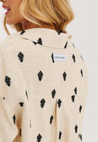 Béžová košile s černě vyšitými diamantovými vzory, s límcem na knoflíky a štítkem s nápisem "BOHO PRERIE" na zadní straně.