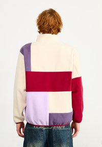 Fleecepullover med høj krave, prydet med et patchwork-design i creme, rød, lavendel og lilla. Blød tekstur med elastisk kant.