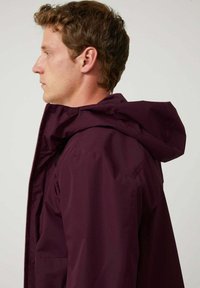 Chaqueta impermeable burdeos con capucha, cremallera visible y bolsillos laterales. Textura suave, forma ajustada y diseño minimalista.