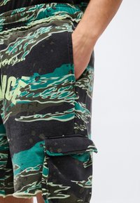 Κοντινή λήψη ενός χεριού μέσα στην τσέπη μαύρου και πράσινου παραλλαγμένου σορτς cargo με τσέπη με καπάκι στο πόδι και μερικό κείμενο ορατό.