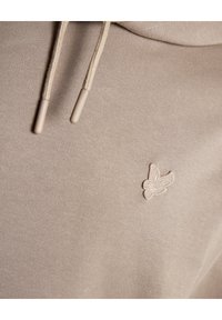 Beige hoodie tyg med två dragsnören och en liten broderad flygande fågel detalj på bröstområdet.