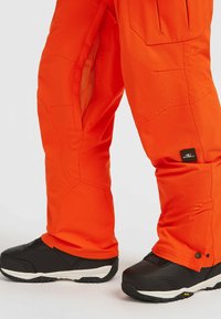 Heldere oranje skipantalons met een versterkt kniegedeelte, ritszakken aan de zijkant en zwarte, rubber-soled laarzen met witte accenten.