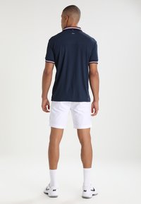 Marineblauw polo shirt met rood en wit gestreepte kraag, korte mouwen, gecombineerd met witte shorts. Bevat een logo bij de hals en heeft een strakke uitstraling.