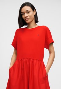 Κόκκινο φόρεμα με στρογγυλή λαιμόκοψη, κοντά μανίκια, φούστα με πιέτες και πλαϊνές τσέπες. Κατασκευασμένο από απαλό, ελαφρύ ύφασμα.