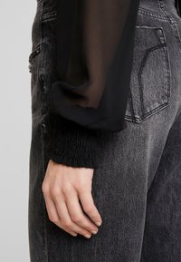 Svart genomskinlig blus med långa ärmar och detaljerade smockade manschetter, matchad med urtvättade, mörkgrå, hög midja jeans med slitna detaljer.