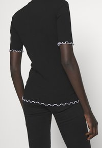 Persona vistiendo un top negro ajustado de manga corta con bordes rizados blancos en las mangas y el dobladillo, combinado con pantalones negros, vista desde atrás.