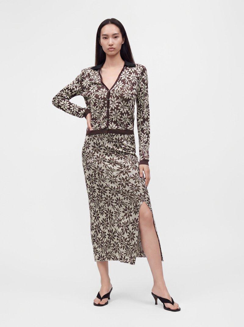 Le mannequin pose dans un haut boutonné marron et crème à motif floral et une jupe midi assortie avec une fente latérale, portant des talons noirs sur un fond blanc.
