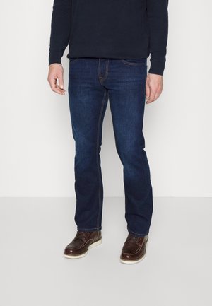 Hombre vestido con vaqueros azul oscuro, suéter oscuro y zapatos de cuero marrón, de pie frente a un fondo claro y liso.