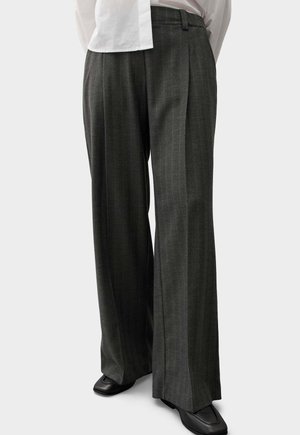 Personne portant un pantalon large gris foncé à fines rayures, une chemise blanche rentrée et des chaussures noires à bout carré sur un fond clair.