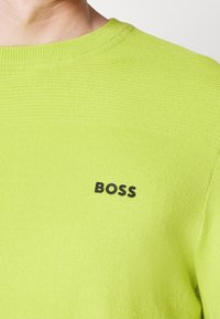 Närbild av en person som bär en limegrön stickad tröja med "BOSS" tryckt i svart på bröstet.