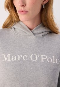 Hellgraue Kapuzenjacke aus weichem Material, mit einem großen weißen Aufdruck von "Marc O'Polo" auf der Vorderseite und einer Kordelzugkapuze.