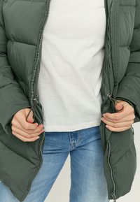 Puffer jacket in dark green fabric with a zippered front, featuring a quilted design. Paired with a white shirt and blue jeans.