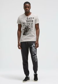 Hellgraues T-Shirt mit Grafikdruck und schwarzem Text, kombiniert mit schwarzen Jogginghosen mit weißen Seitenstreifen und bedruckten Logos.