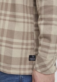 Bruine en beige geruite overhemd van katoen, met een label aan de zijkant en zichtbare stiksels langs de naden.