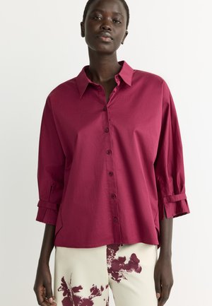 Woman wearing a loose-fitted dark pink button-up blouse with 3/4 sleeves and white pants with dark pink floral patterns.