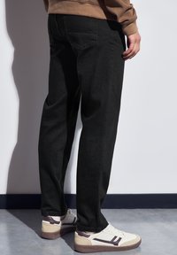 Pantalones de mezclilla negros con un diseño de pierna recta, que cuentan con bolsillos traseros y un acabado texturizado, combinados con zapatillas crema y marrón.