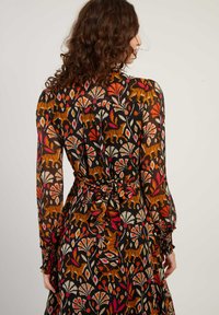 Vrouw met krullend haar die een lange jurk draagt met een oranje, rode en beige bloemen- en tijgerprint, die in de taille is gestrikt, met haar rug naar ons toegekeerd.