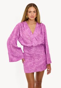 Robe mini violette en tissu texturé, avec décolleté en V et manches longues évasées. Elle présente une taille cintrée et des détails en couches.