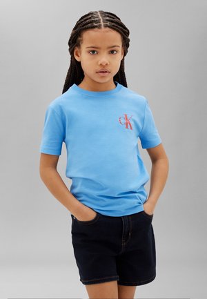 Młoda osoba z zaplecionymi warkoczami, ubrana w jasnoniebieski T-shirt Calvin Klein i czarne szorty, z rękami w kieszeniach, stojąca na szarym tle.