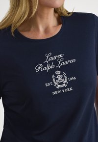 Γυναίκα που φοράει μπλε πουκάμισο με λευκό κεντημένο κείμενο "Lauren Ralph Lauren," ένα έμβλημα και "EST. 1996 NEW YORK" στο στήθος.