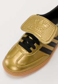Zlaté syntetické tenisky s hladkým povrchem, černými tkaničkami, třemi černými pruhy a přesměrně velkým zlatým jazykem s logem Adidas.