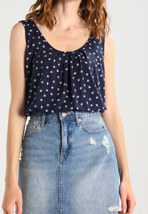 Kvinde iført en ærmeløs navy top med små hvide print, stoppet ned i et lyseblåt distressed denimskørt.