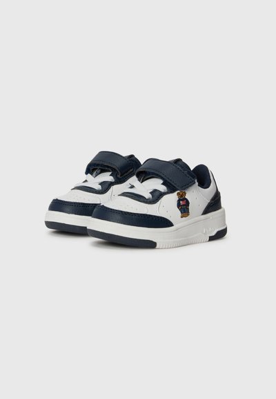 Zapatillas blancas y navy con una superficie de cuero texturizado, que presentan un emblema de oso, correas de velcro y una suela de goma.