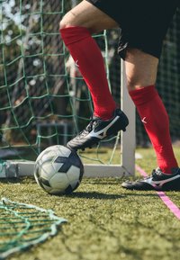 Saboți de fotbal negri cu accente argintii pe un teren de iarbă, șosete roșii și o minge de fotbal alb-negru așezată sub picior.