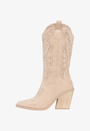 Bota cowboy de ante beige con punta afilada y patrones de costura curvados. Presenta un tacón de madera gruesa y texturizada, y lengüetas para facilitar su uso.