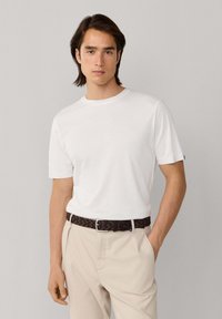 Homme aux cheveux foncés de longueur moyenne, portant une chemise blanche à manches courtes, un pantalon beige et une ceinture tressée foncée, debout devant un fond clair et uni.