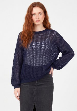 Giovane donna con lunghi capelli rossi che indossa un maglione blu navy a maglia aperta sopra una canottiera scura e una gonna scura con uno spacco frontale.