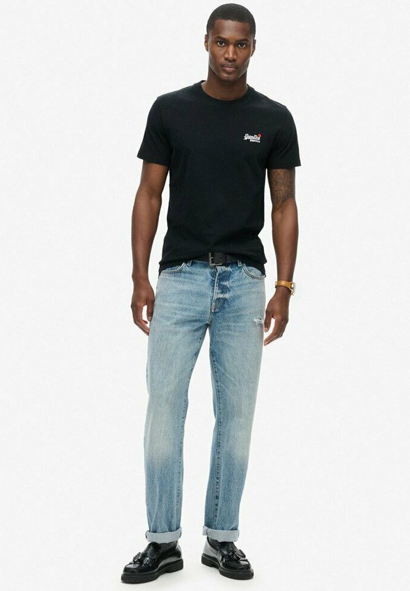 Camiseta de algodón negra de manga corta con un pequeño logo, combinada con jeans de denim desgastados de color azul claro y zapatos de cuero negros.