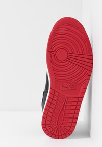 Zapatilla negra con parte superior texturizada, que presenta una suela roja de goma con patrones circulares y un logo central.