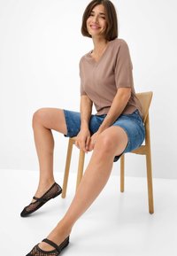 Top marrone a maniche corte con scollo a V, abbinato a pantaloncini di jeans blu. Ballerine nere in rete su una sedia di legno con sfondo semplice.