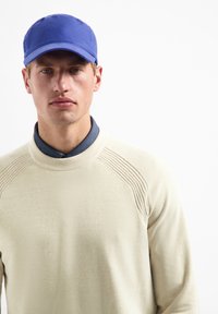 Beige structuurtrui met geribbelde halslijn en schouderdetails, gecombineerd met een effen blauwe pet. Het ontwerp is casual en modern.