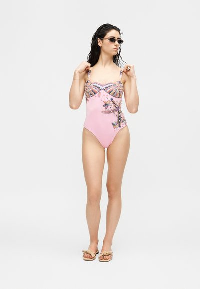 Femme portant un maillot de bain une pièce rose avec un motif coloré d'étoiles de mer, des lunettes de soleil noires et des sandales beige, debout sur un fond blanc.