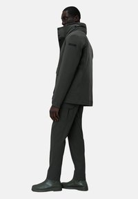 Outerwear verde scuro con colletto alto, tasche laterali e una texture liscia, abbinato a pantaloni cargo coordinati e stivali.