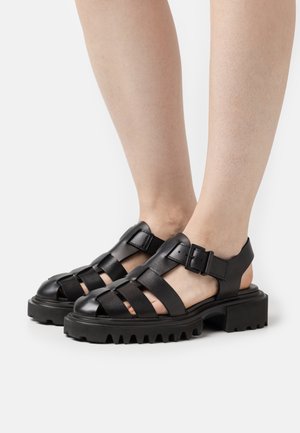 Sorte lædersandaler med flere krydsede stropper, en justerbar spænde og en chunky tekstureret sål. Åben hæl-design for åndbarhed.