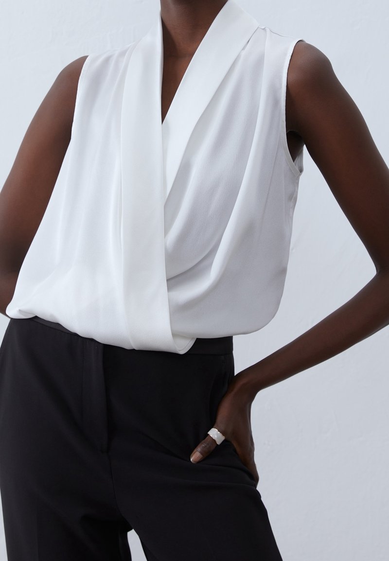 Blouse blanche sans manches avec col en V drapé, associée à un pantalon noir taille haute, la main du mannequin reposant sur la hanche porte une bague blanche.