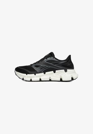 Zapatilla de running negra con parte superior de malla texturizada, detalles en gris y una suela gruesa blanca con detalles recortados. Marca contorneada visible.