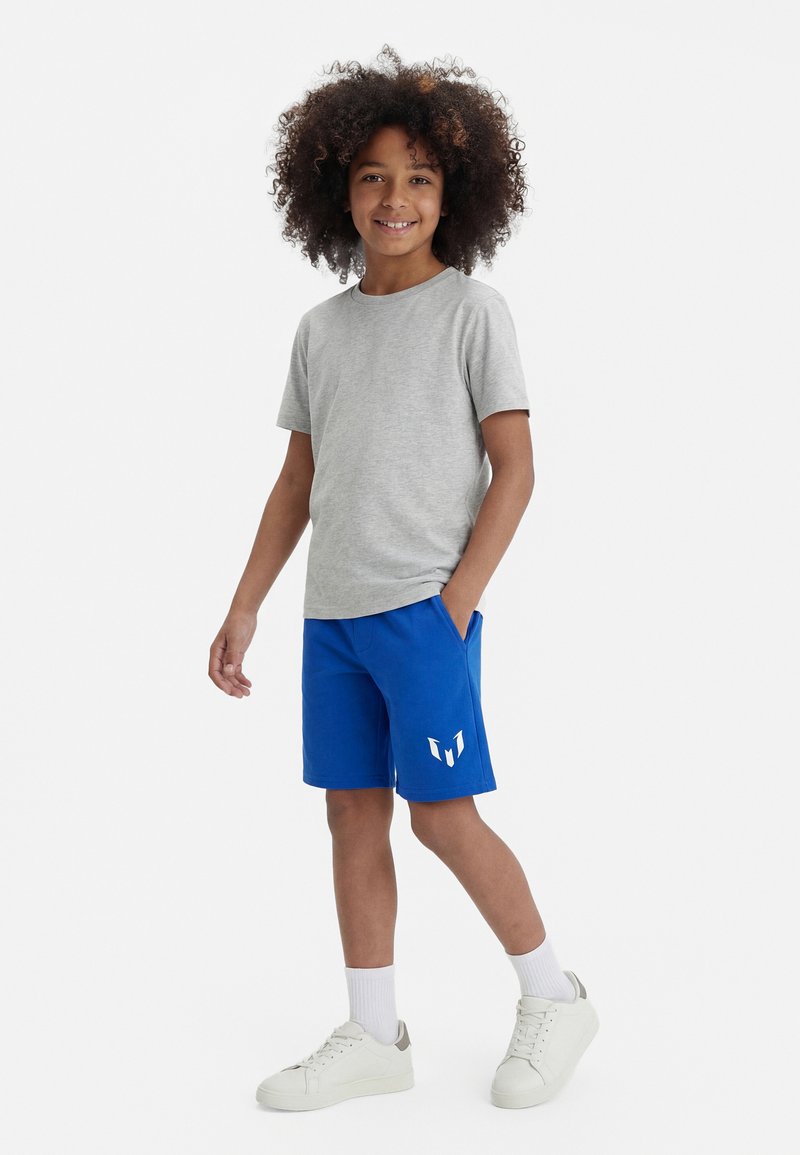 Enfant aux cheveux bouclés portant un t-shirt gris, un short bleu avec un logo blanc, des chaussettes blanches et des baskets blanches, debout avec une main dans la poche.