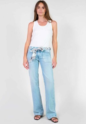 Jeans a zampa d'elefante di colore blu chiaro con una cintura anteriore in tessuto a fantasia e tasche frontali, abbinati a una canotta bianca e sandali neri.