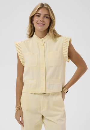 Ung kvinde med blondt hår smiler iført en lysegul ærmeløs bluse med flæser og matchende bukser, med guldsmykker og ur.