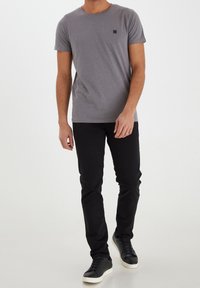 Šedé tričko s krátkým rukávem, kulatým výstřihem a malým černým logem na hrudi, spárované s černými slim-fit kalhotami a černými teniskami.