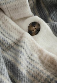 Tkanina z mieszanki wełny w kremowym, szarym i niebieskim wzorze w kratę, z ciemnym guzikiem z czterema otworami i gładką teksturą.