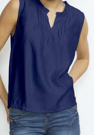 Blouse sans manches bleu marine avec un col en V fendu et de subtils plis verticaux, portée avec un jean en denim bleu. Mains partiellement dans les poches.